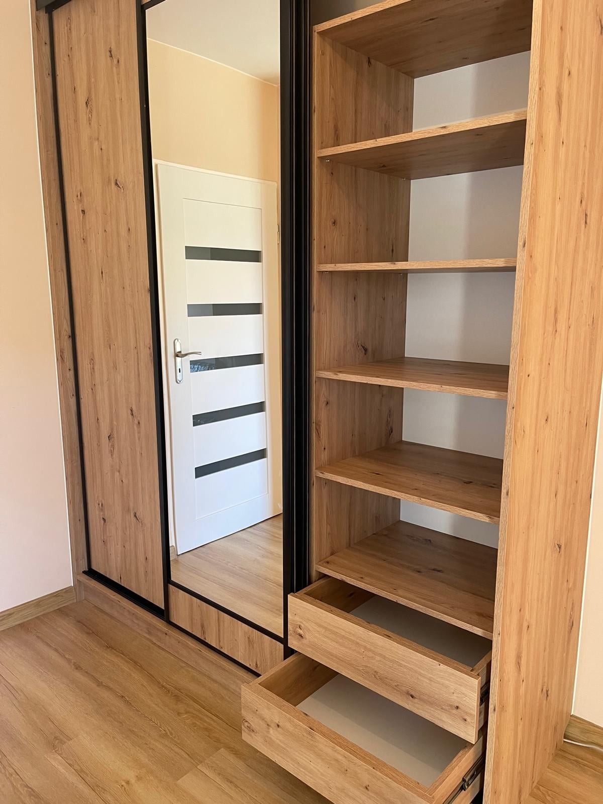 Drewniana szafa z przesuwnymi drzwiami z lustrem, otwartymi półkami i wysuniętymi szufladami, widoczna w odbiciu biała drzwi z czarnymi paskami.