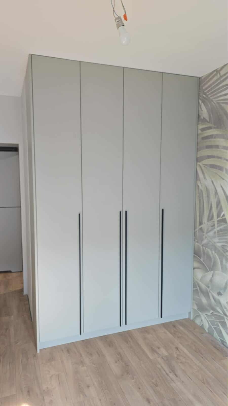 Jasnoszara szafa z czterema drzwiami i długimi, czarnymi uchwytami, na tle ściany z motywem liści. Podłoga z paneli imitujących drewno. Widoczna żarówka bez klosza.