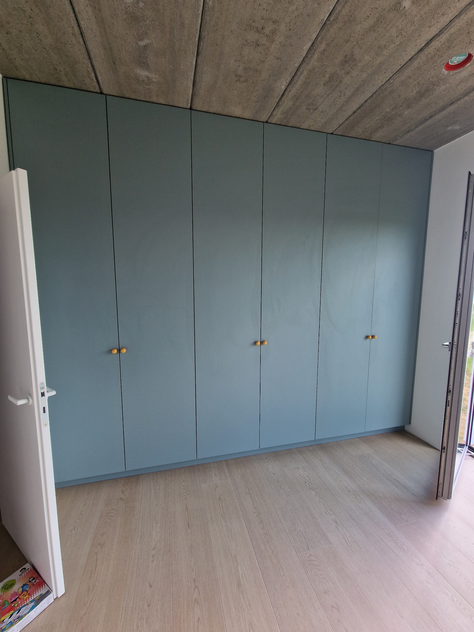 Błękitna szafa wnękowa z drewnianymi uchwytami, w minimalistycznym wnętrzu z betonowym sufitem i jasną podłogą. Widok na otwarte drzwi balkonowe.