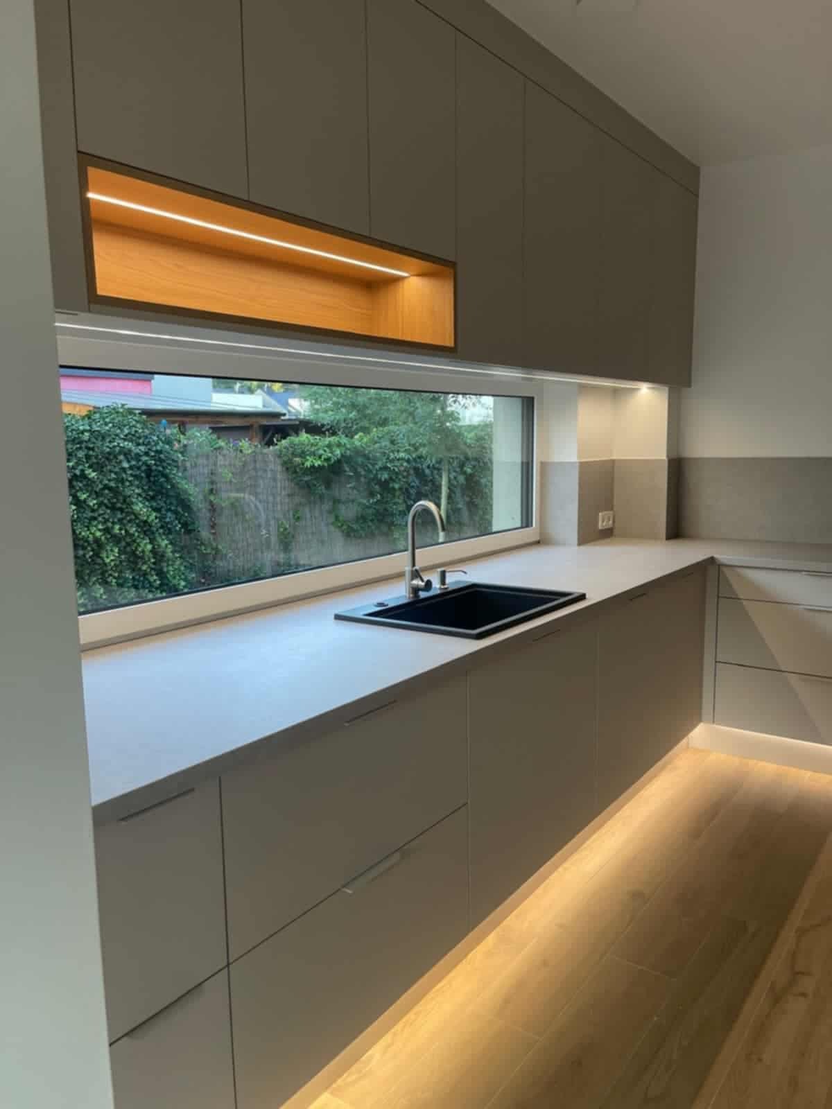 Nowoczesna kuchnia z szarymi szafkami, czarnym zlewem i oknem z widokiem na zieleń. Podświetlenie LED pod szafkami i w górnej wnęce dodaje ciepła. Minimalistyczny design.
