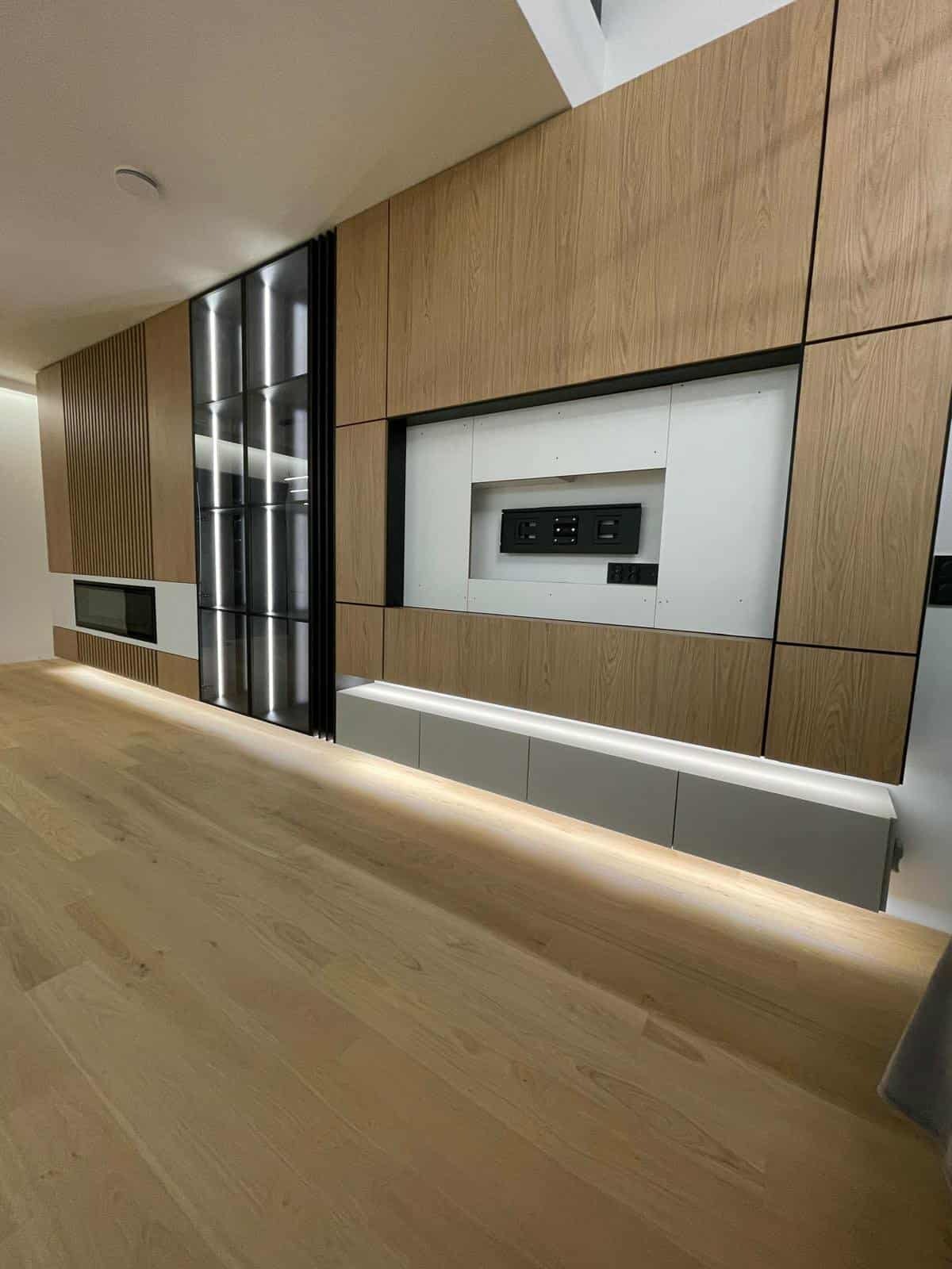 Drewniana zabudowa ściany z szafkami, wnęką na TV i przeszkloną witryną z podświetleniem LED. Nowoczesny design, jasne drewno, minimalistyczny styl.