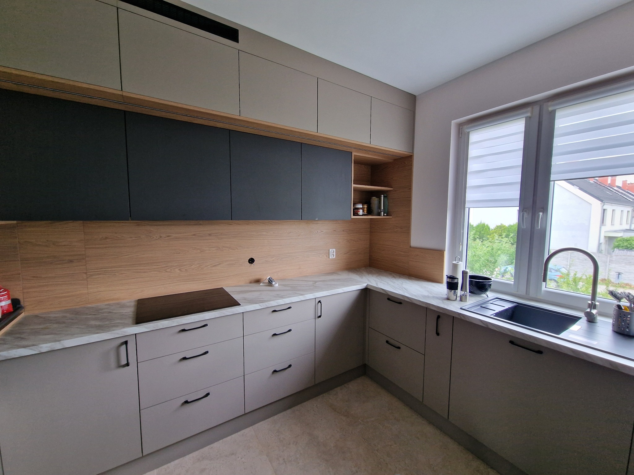 Jasna kuchnia na wymiar z drewnianym panelem ściennym i czarnymi frontami szafek górnych. Widoczny zlew pod oknem i płyta indukcyjna. Minimalistyczny design.