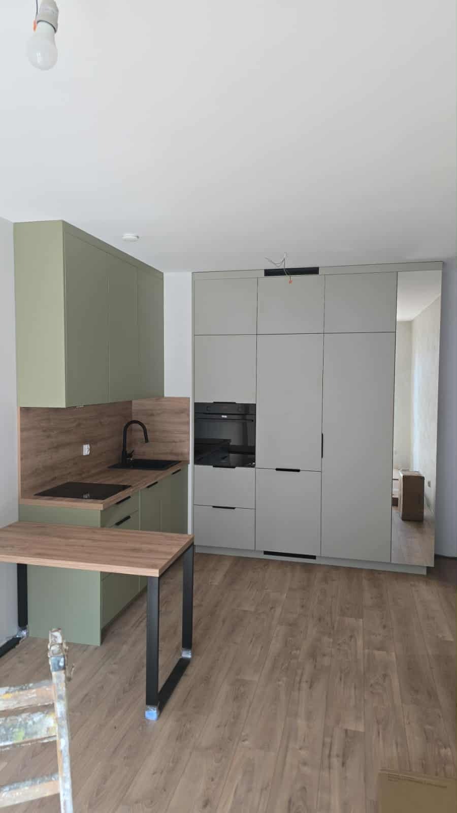 Nowoczesna kuchnia z zielonymi szafkami i szarą zabudową z piekarnikiem, uzupełniona drewnianym blatem stołu na metalowych nogach, na tle drewnianej podłogi.