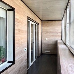 Builder - Wnętrze balkonu wykończonego panelami ściennymi imitującymi drewno, z czarną podłogą i białymi ramami okiennymi oraz drzwiami balkonowymi.