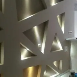 Ściana z gładzi z geometrycznymi wnękami oświetlonymi punktowo, widoczne puszki farb przy podłodze.