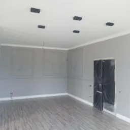 Szare, odnowione wnętrze z panelami podłogowymi, ozdobnymi listwami i przygotowaną do malowania ścianą z czarnymi, minimalistycznymi lampami sufitowymi i zabezpieczonymi folią drzwiami.