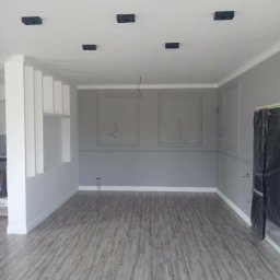 Builder - Szare wnętrze w Krakowie, z panelami podłogowymi imitującymi drewno, szare ściany z białymi listwami i czarnymi lampami sufitowymi, w trakcie prac wykończeniowych.