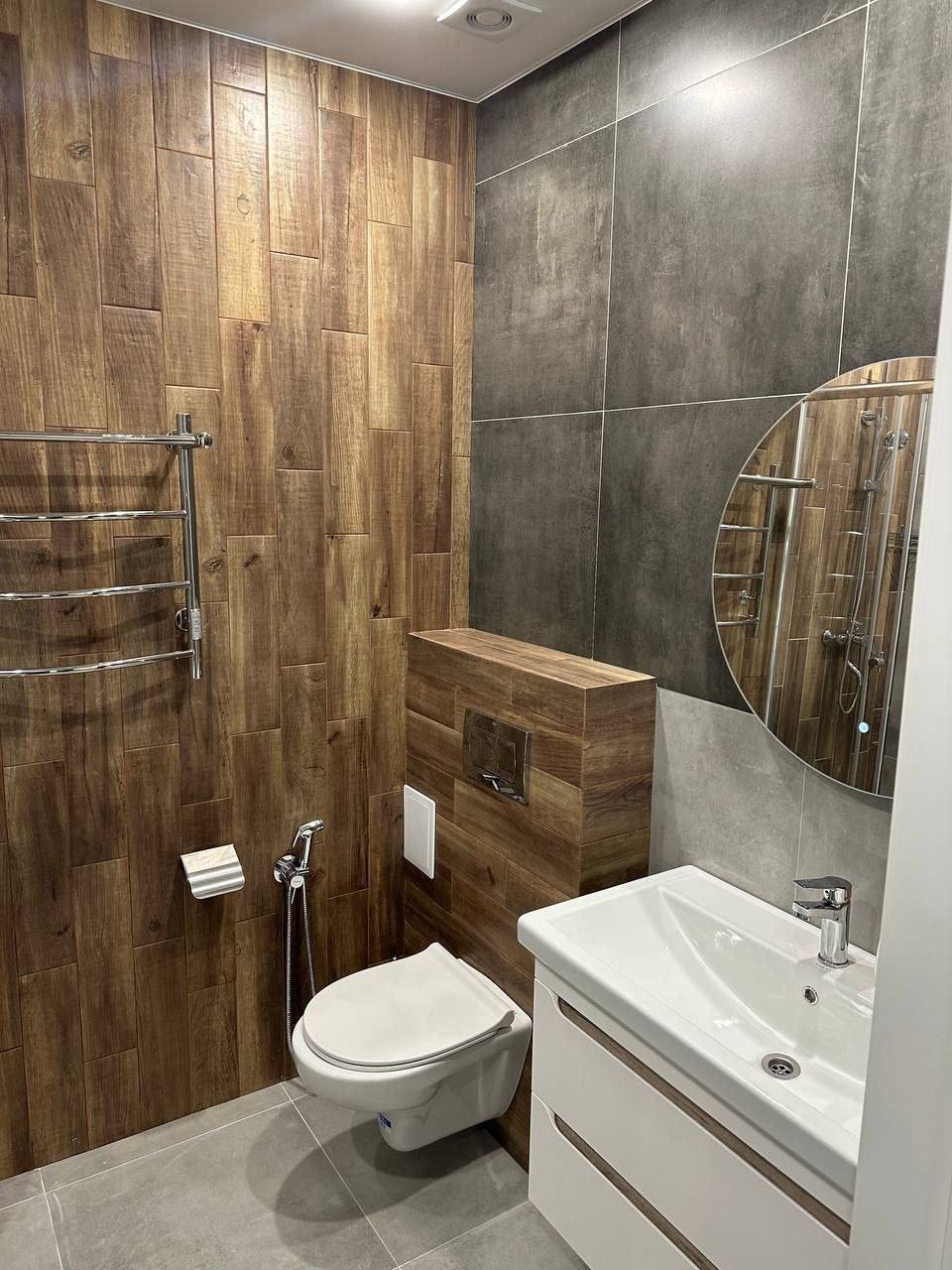 Nowoczesna łazienka z drewnopodobnymi płytkami na ścianie, wiszącą toaletą z bidetką, okrągłym lustrem i szarą płytką na ścianie obok umywalki.