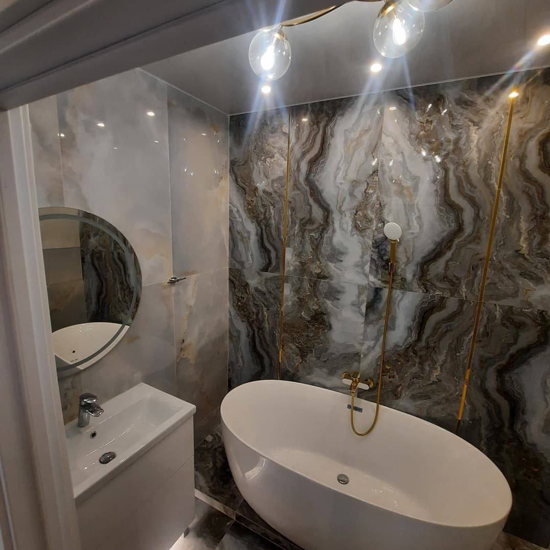 Elegancka łazienka w Krakowie z owalną wanną, marmurowymi płytkami na ścianach i okrągłym lustrem nad umywalką, oświetlona lampą sufitową z kulistymi kloszami.