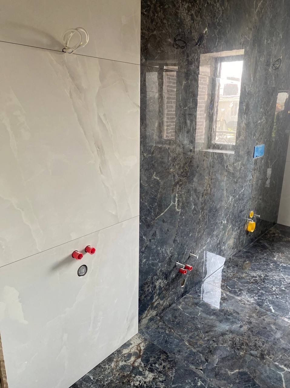 Łazienka w trakcie remontu, widoczne dwie ściany wyłożone płytkami imitującymi marmur w kolorach szarości i bieli, z widocznymi instalacjami wodnymi i elektrycznymi.