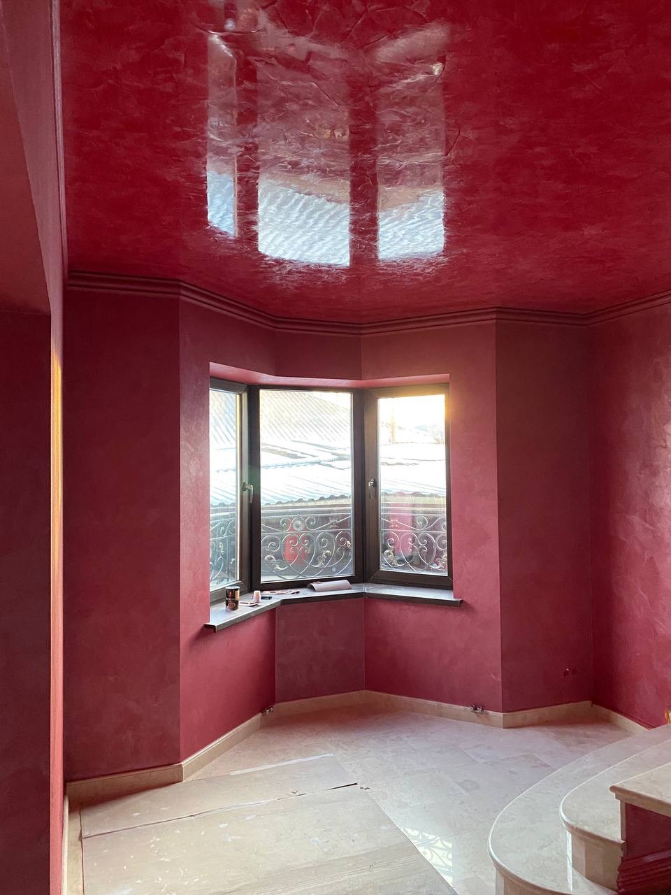 Pokój po remoncie z czerwonymi ścianami i sufitem o błyszczącej, marmurkowatej fakturze, widoczne okno narożne z ozdobną kratą, podłoga częściowo zabezpieczona kartonem.