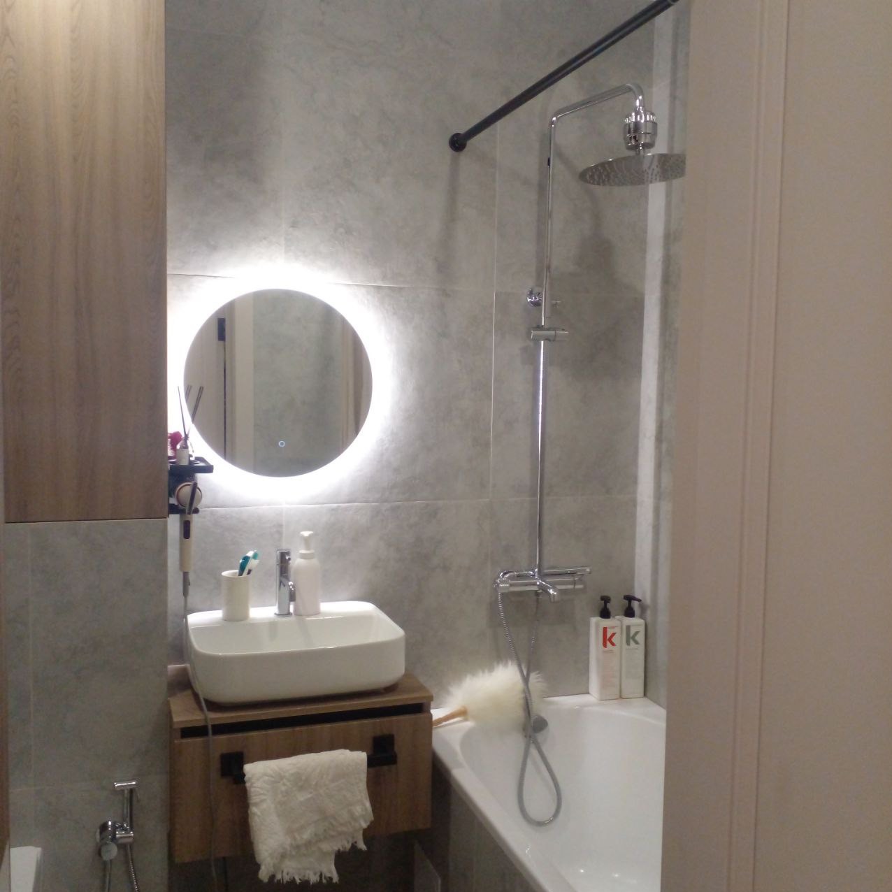 Nowoczesna łazienka z okrągłym lustrem podświetlanym LED, białą umywalką nablatową na drewnianej szafce z czarnymi uchwytami, szarą glazurą i chromowaną baterią prysznicową.