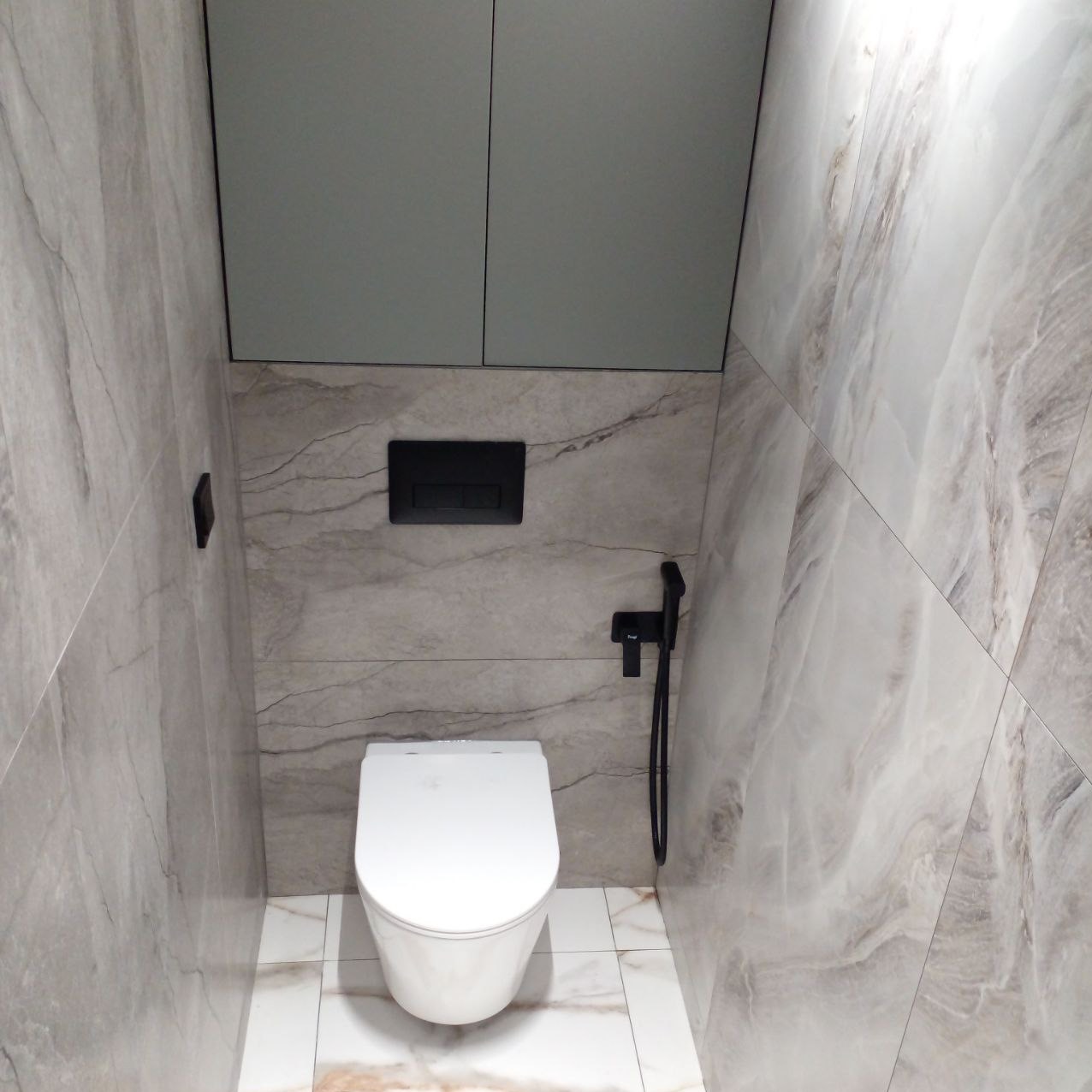 Nowoczesna toaleta podwieszana z czarną armaturą, obudowana płytami imitującymi marmur, z zabudową szafkową nad stelażem.