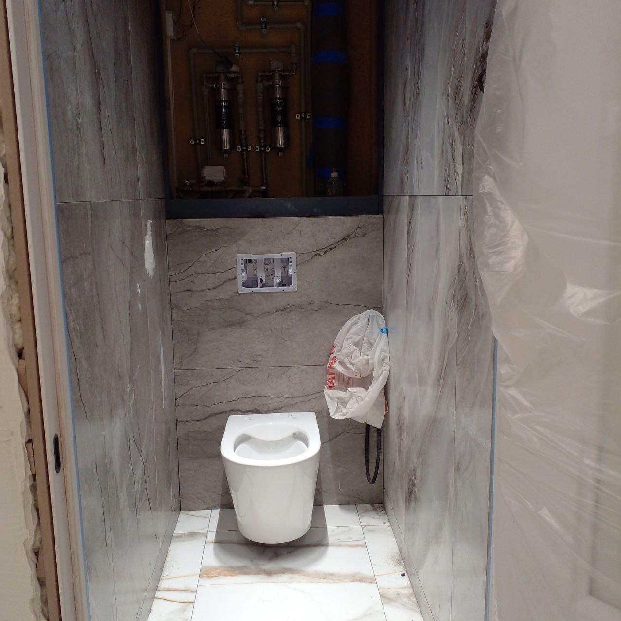 Wanna wisząca zamontowana w wąskiej, wykończonej szarą płytką łazience, widoczna instalacja wodna nad zabudową, biała torba foliowa wisząca na ścianie, częściowo zakryta folią ochronną.