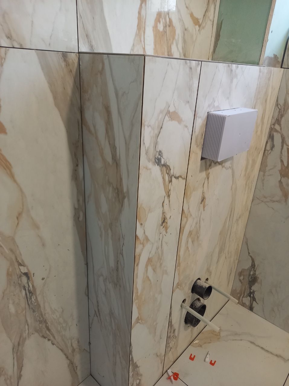 Ściana i podłoga wyłożone płytkami imitującymi marmur w odcieniach beżu i brązu. Widoczne otwory na rury i biały element wentylacyjny na ścianie, a także czerwone krzyżyki dystansowe na podłodze.