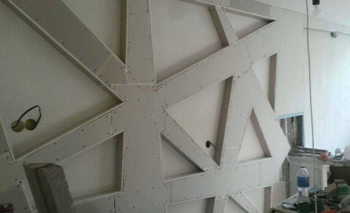 Nierówna ściana z płyt kartonowo-gipsowych tworzących geometryczne wzory, widoczne kable elektryczne, niedokończone wykończenie, butelka wody w tle.