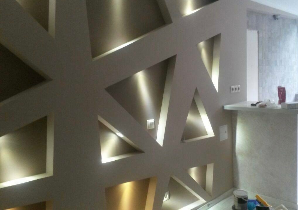 Ściana z gładzi z geometrycznymi wnękami oświetlonymi punktowo, widoczne puszki farb przy podłodze.