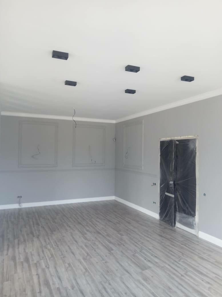 Szare, odnowione wnętrze z panelami podłogowymi, ozdobnymi listwami i przygotowaną do malowania ścianą z czarnymi, minimalistycznymi lampami sufitowymi i zabezpieczonymi folią drzwiami.