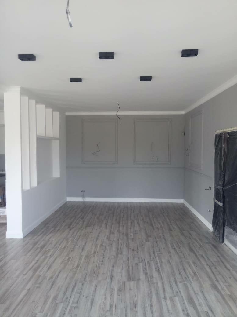 Szare wnętrze w Krakowie, z panelami podłogowymi imitującymi drewno, szare ściany z białymi listwami i czarnymi lampami sufitowymi, w trakcie prac wykończeniowych.