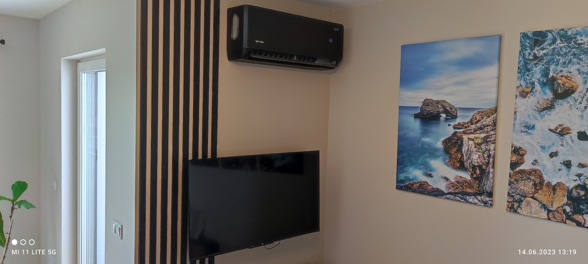 Czarny klimatyzator ścienny z wyświetlaczem temperatury na ścianie w salonie, obok pionowe panele dekoracyjne i obrazy z motywem morskim.