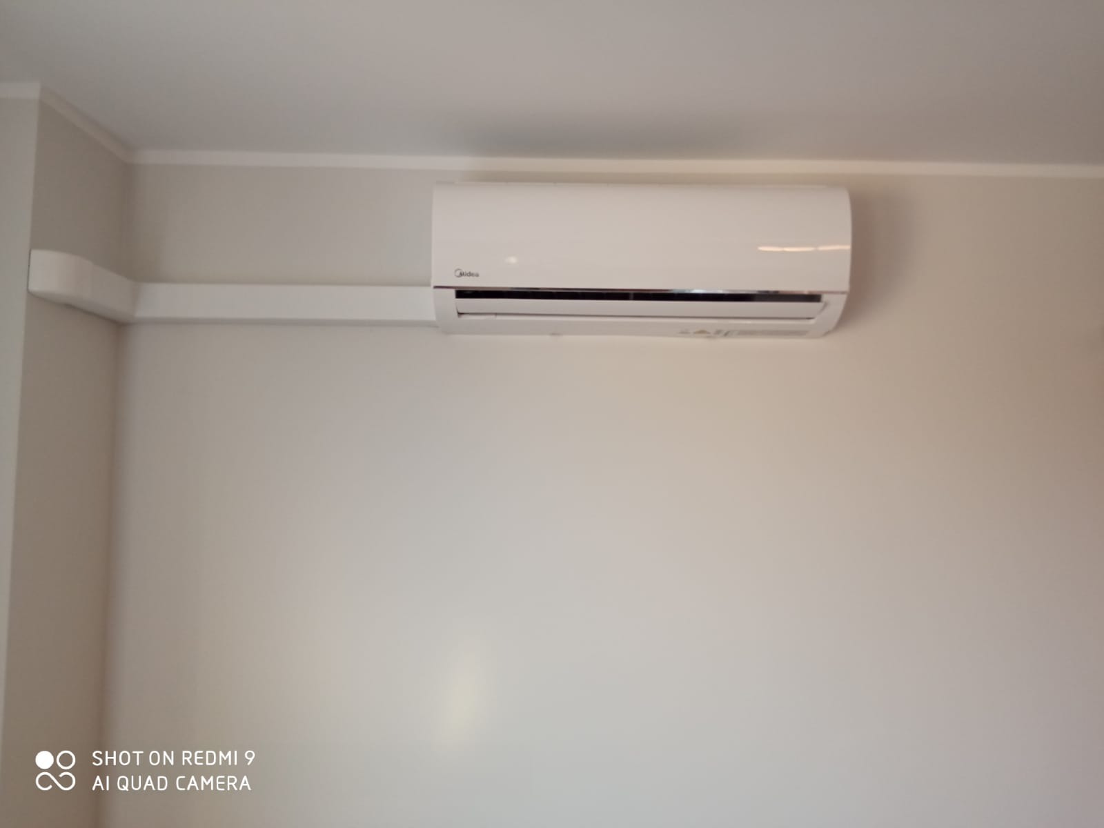 Biała jednostka wewnętrzna klimatyzatora marki Midea zamontowana na jasnej ścianie, z widoczną listwą maskującą przewody.