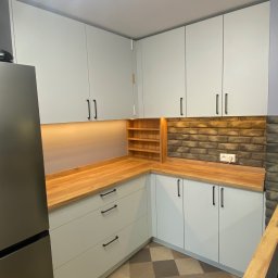 Kacper Stanisławski - Jasna kuchnia z drewnianym blatem, oświetleniem podszafkowym i ceglaną ścianą. Widoczny fragment lodówki i podłoga w szachownicę.