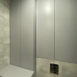 Szare szafki w łazience, zabudowa nad pralką i WC, z widocznymi elementami montażowymi. Minimalistyczny design, jasne płytki ceramiczne w tle.