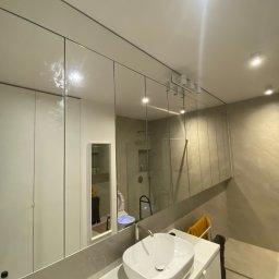 Kacper Stanisławski - Nowoczesna łazienka z szafkami na wymiar z lustrzanymi frontami, odbijającymi wnętrze. Umywalka nablatowa i oświetlenie punktowe. Widoczny fragment osoby robiącej zdjęcie.