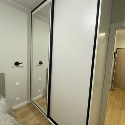 Kacper Stanisławski - Biała szafa przesuwna z czarnymi obramowaniami i lustrem, widoczna w rogu pokoju z drewnianą podłogą. Minimalistyczny design, nowoczesne wykończenie.