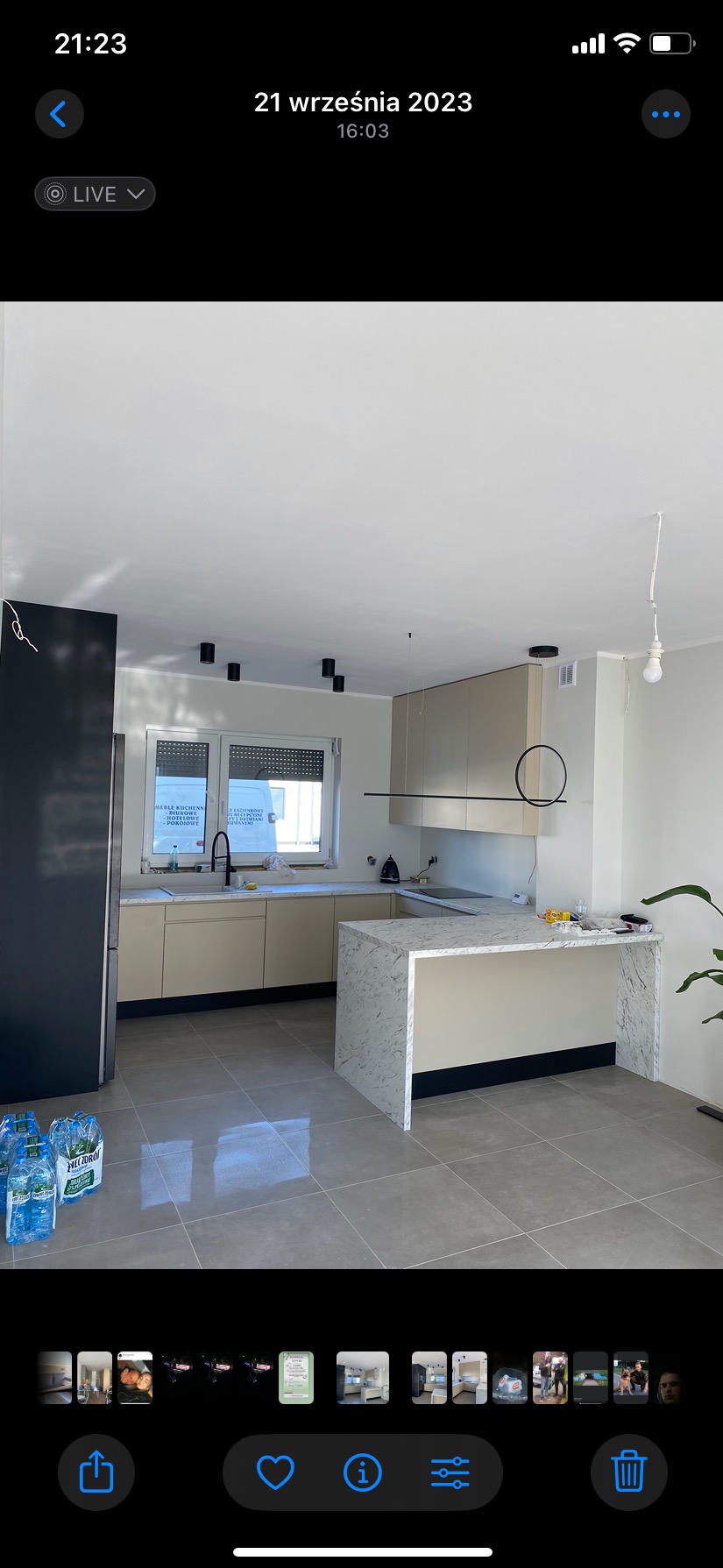 Jasna kuchnia z wyspą wykończoną marmurem, widoczne czarne akcenty w postaci oświetlenia i dolnych paneli, na podłodze szare płytki, butelki z wodą na pierwszym planie.