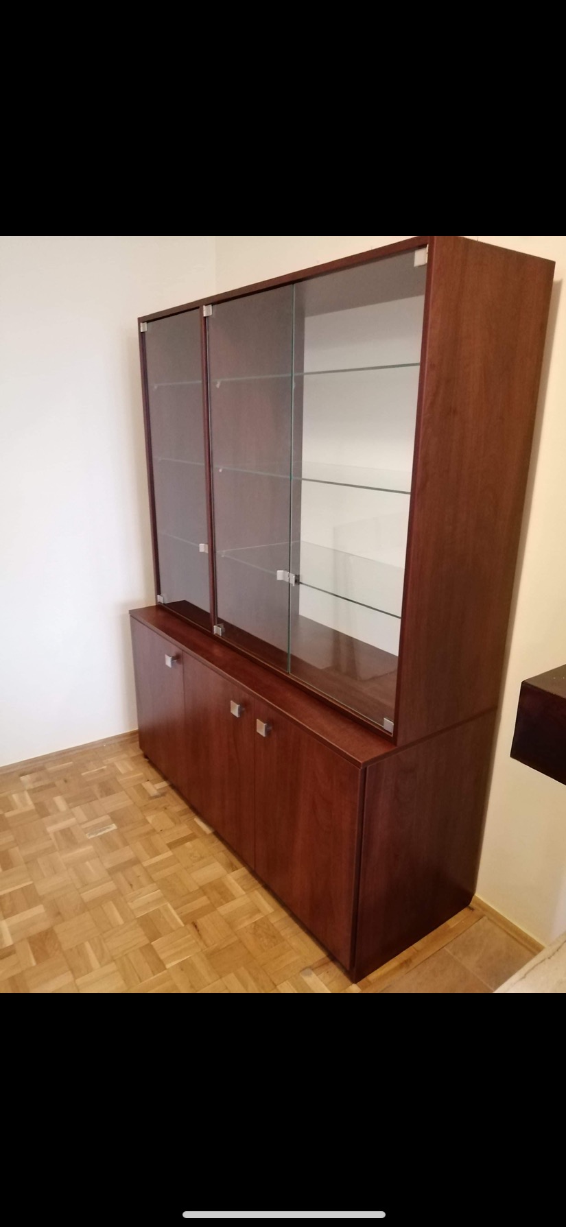 Wysoka, ciemnobrązowa szafa z przeszklonymi drzwiami na górze i pełnymi drzwiami na dole, z metalowymi uchwytami, stojąca na parkiecie w jasnym pomieszczeniu.