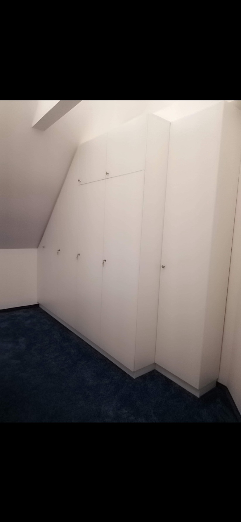 Biała szafa wnękowa na poddaszu z ciemnoniebieską wykładziną podłogową, widoczne srebrne uchwyty i klucz w zamku.