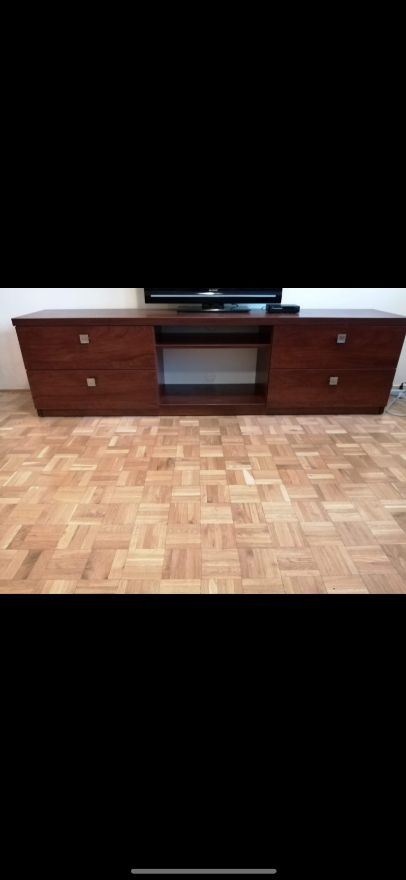 Długa, niska komoda RTV z ciemnego drewna z szufladami i otwartymi półkami, na parkiecie w jodełkę, z telewizorem na górze.