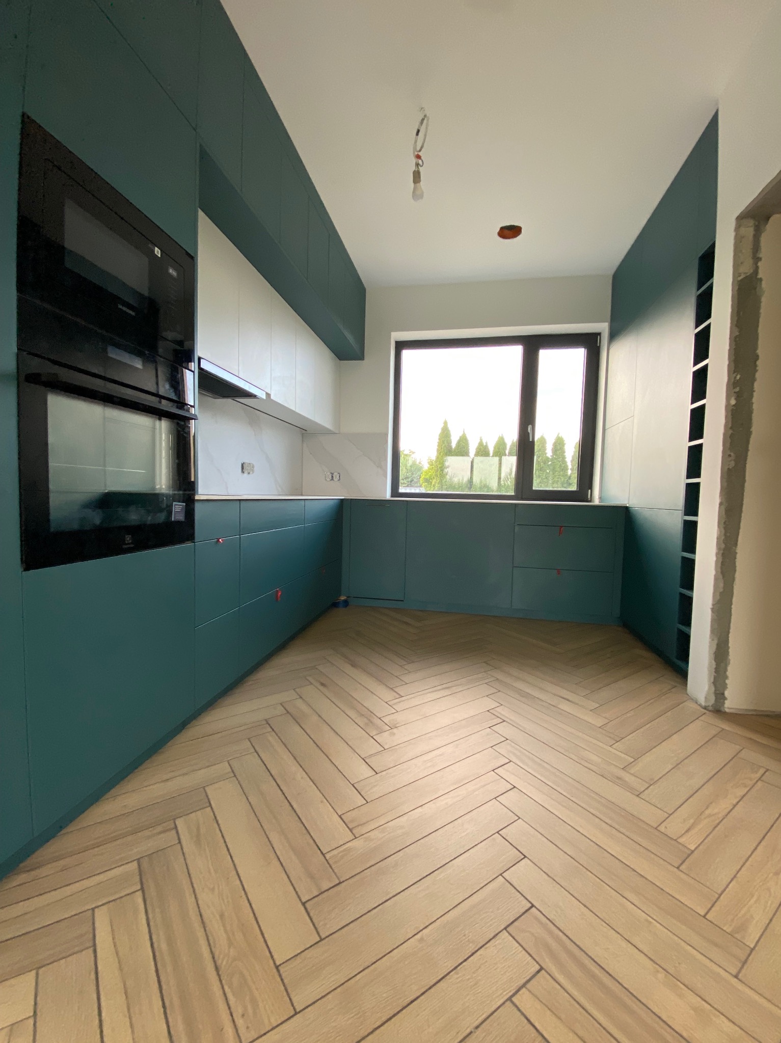 Nowoczesna kuchnia z turkusowymi szafkami i parkietem w jodełkę. Widok na okno z zielenią. Czyste linie, minimalistyczny design.