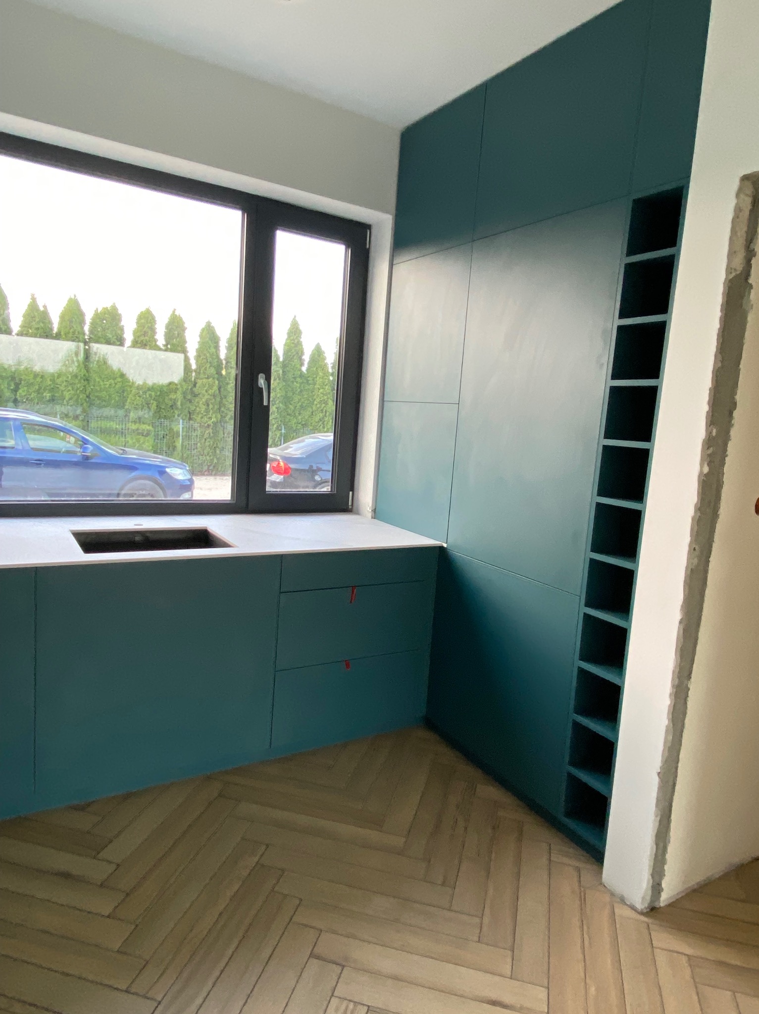 Turkusowa zabudowa kuchenna z szafkami, blatem z otworem na zlew i regałem wnękowym. Parkiet ułożony w jodełkę. Widok na zieleń i auta za oknem.