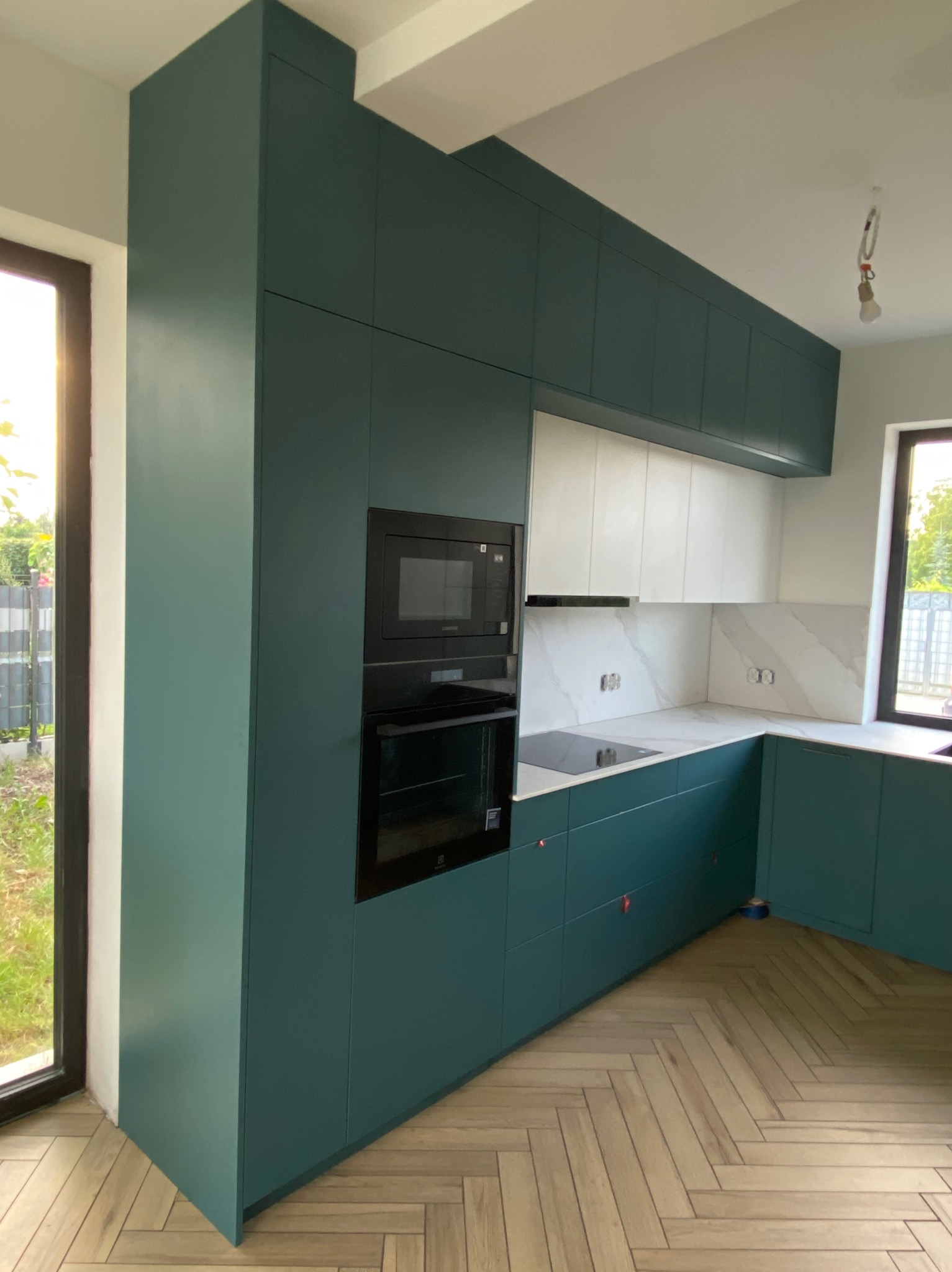 Nowoczesna kuchnia z zielonymi szafkami, białym blatem i marmurową ścianą. Widoczny piekarnik, mikrofalówka i płyta indukcyjna. Podłoga w jodełkę.