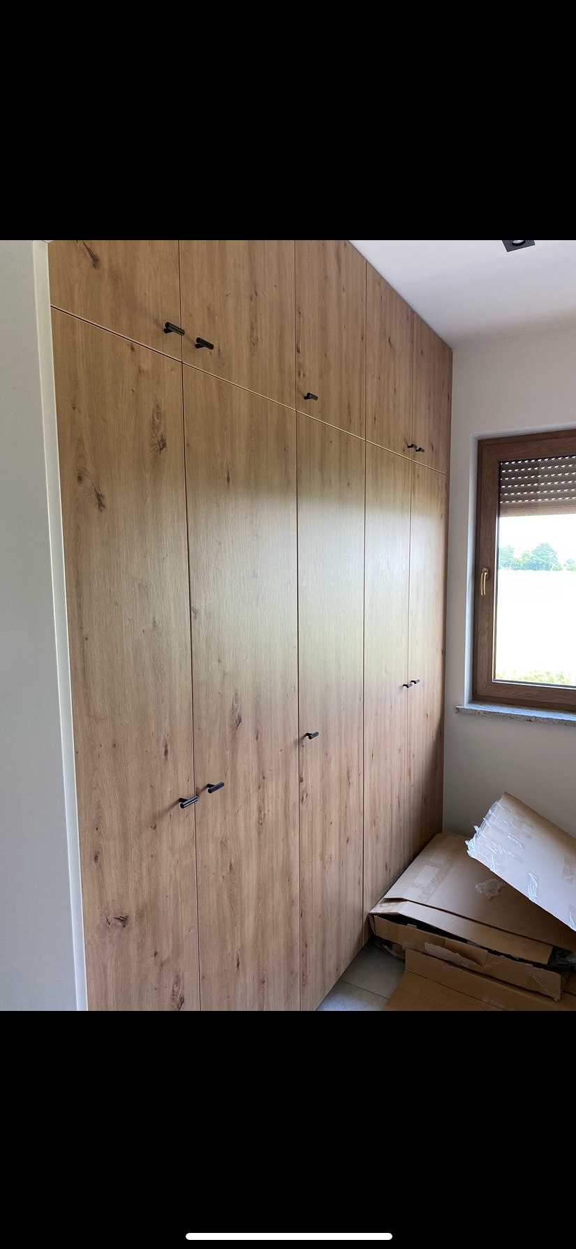 Szafa wnękowa z drewnianymi frontami i czarnymi uchwytami, sięgająca sufitu, z oknem i kartonami w tle, w jasnym wnętrzu.