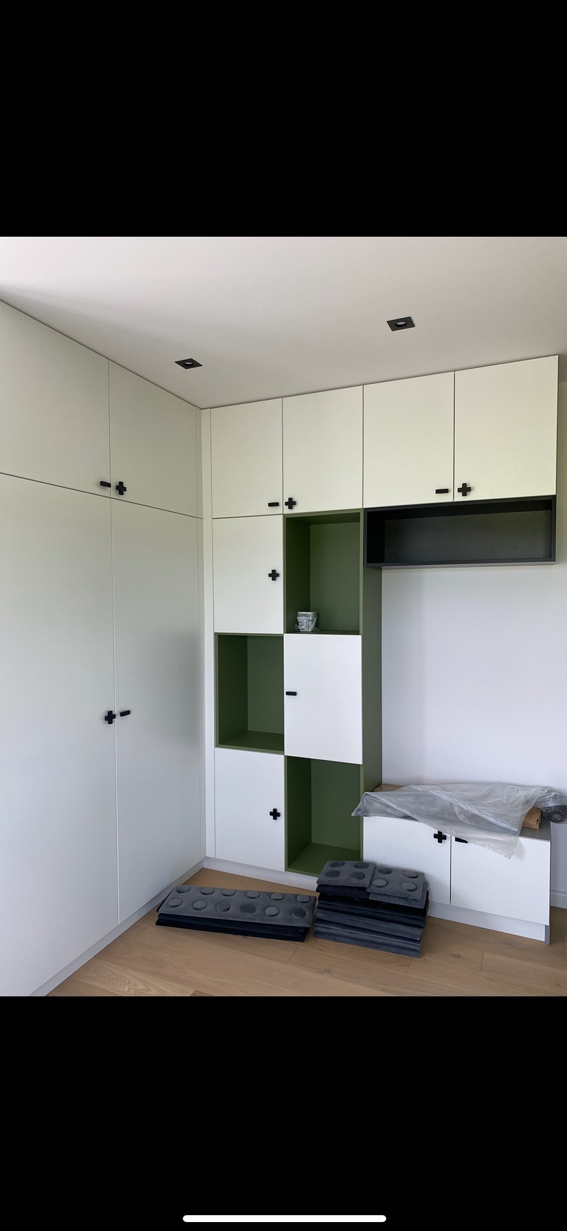 Biała szafa wnękowa z czarnymi uchwytami krzyżowymi i zielonym wnętrzem półek, na podłodze leżą szare poduszki w kształcie klocków Lego.