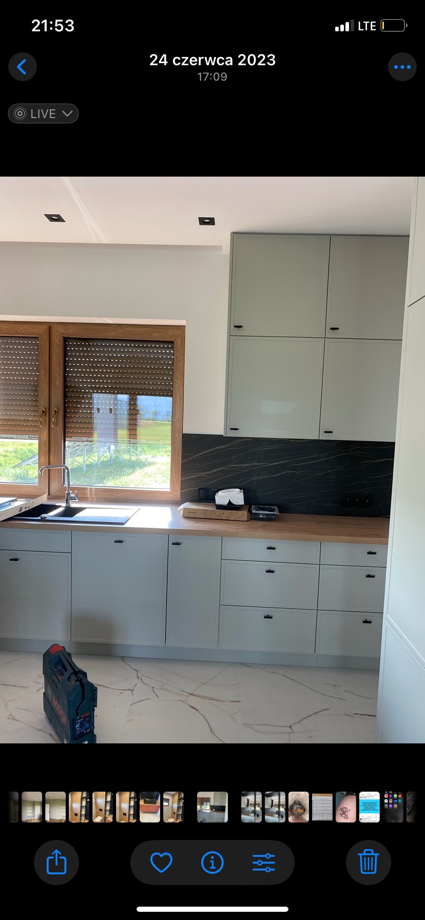 Jasna kuchnia z drewnianym blatem i czarnym panelem nad nim. Na podłodze marmurowe płytki. Przy oknie widoczny drewniany rolety. Na podłodze leży narzędzie.