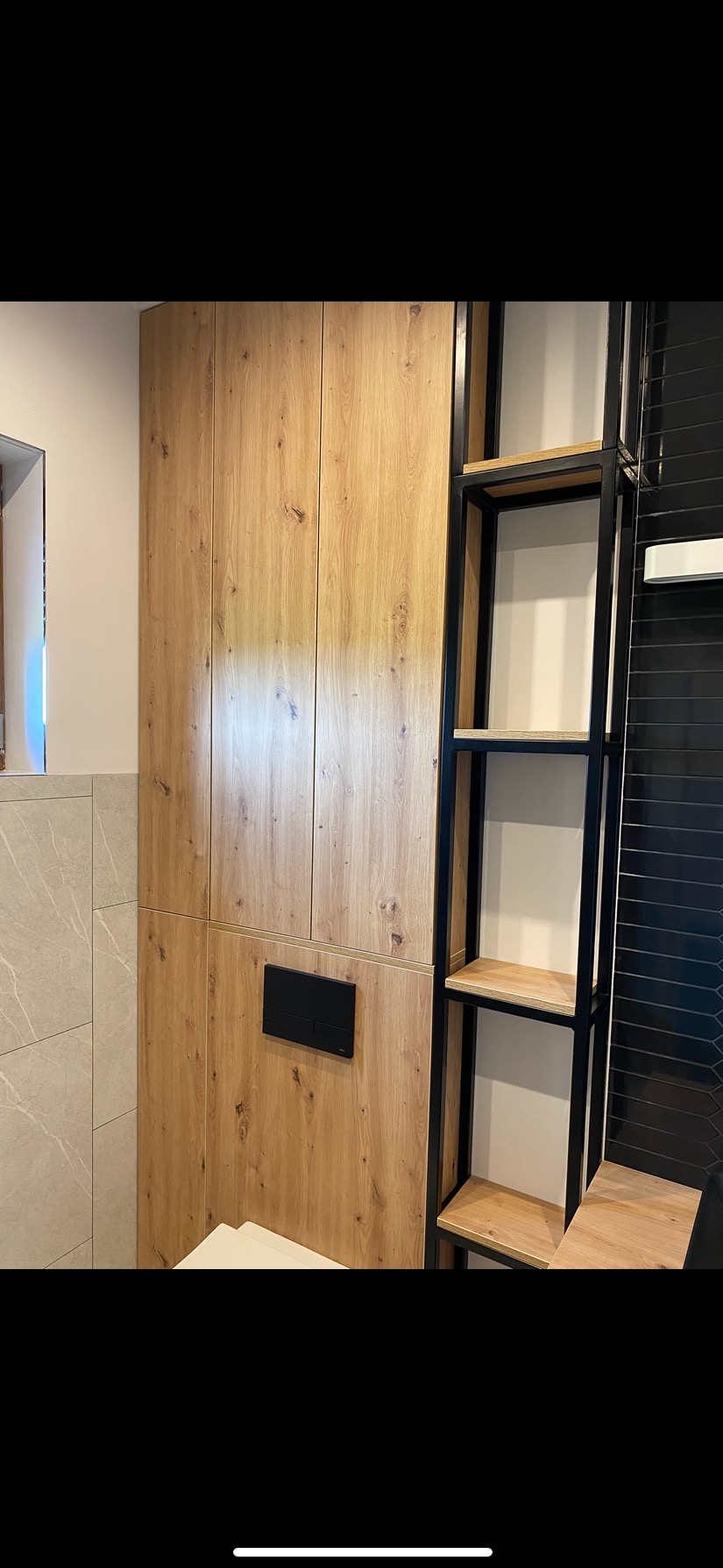 Drewniana szafa na wymiar z czarnym przyciskiem spłuczki i metalowy regał w łazience. Widoczne płytki i fragment toalety.