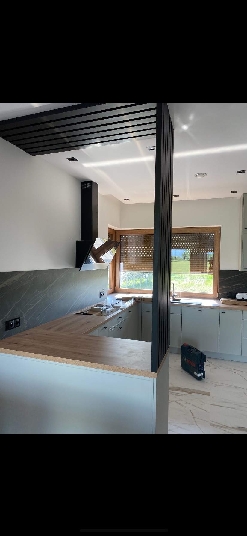 Nowoczesna kuchnia z szarymi szafkami, drewnianym blatem i czarną kolumną dekoracyjną. Widoczny okap i okno z roletami. Na podłodze marmurowe płytki.