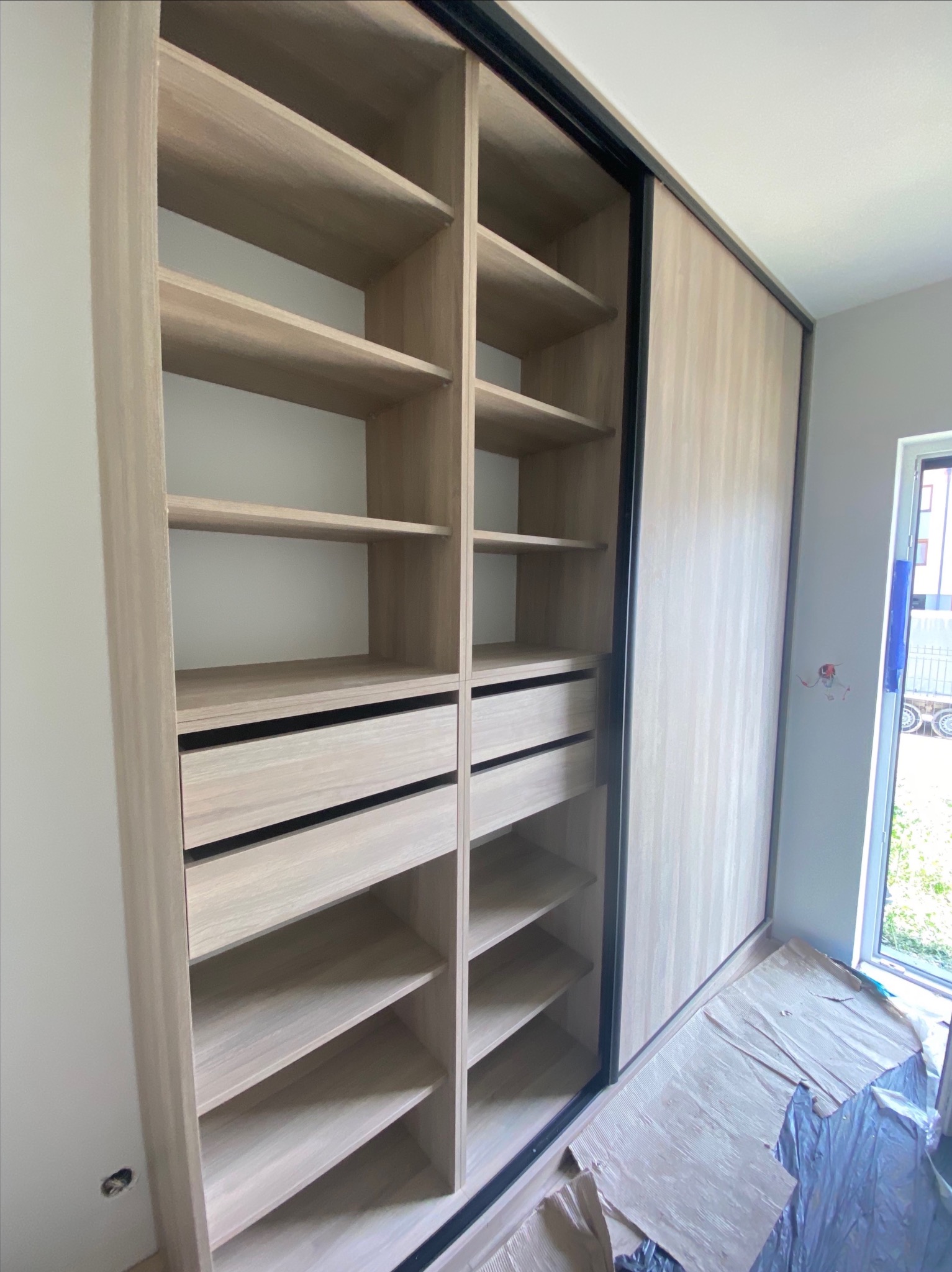 Jasna szafa wnękowa z przesuwnymi drzwiami i półkami. Widoczne szuflady i czarne obramowanie drzwi. Fragment okna w tle, zabezpieczenia na podłodze.