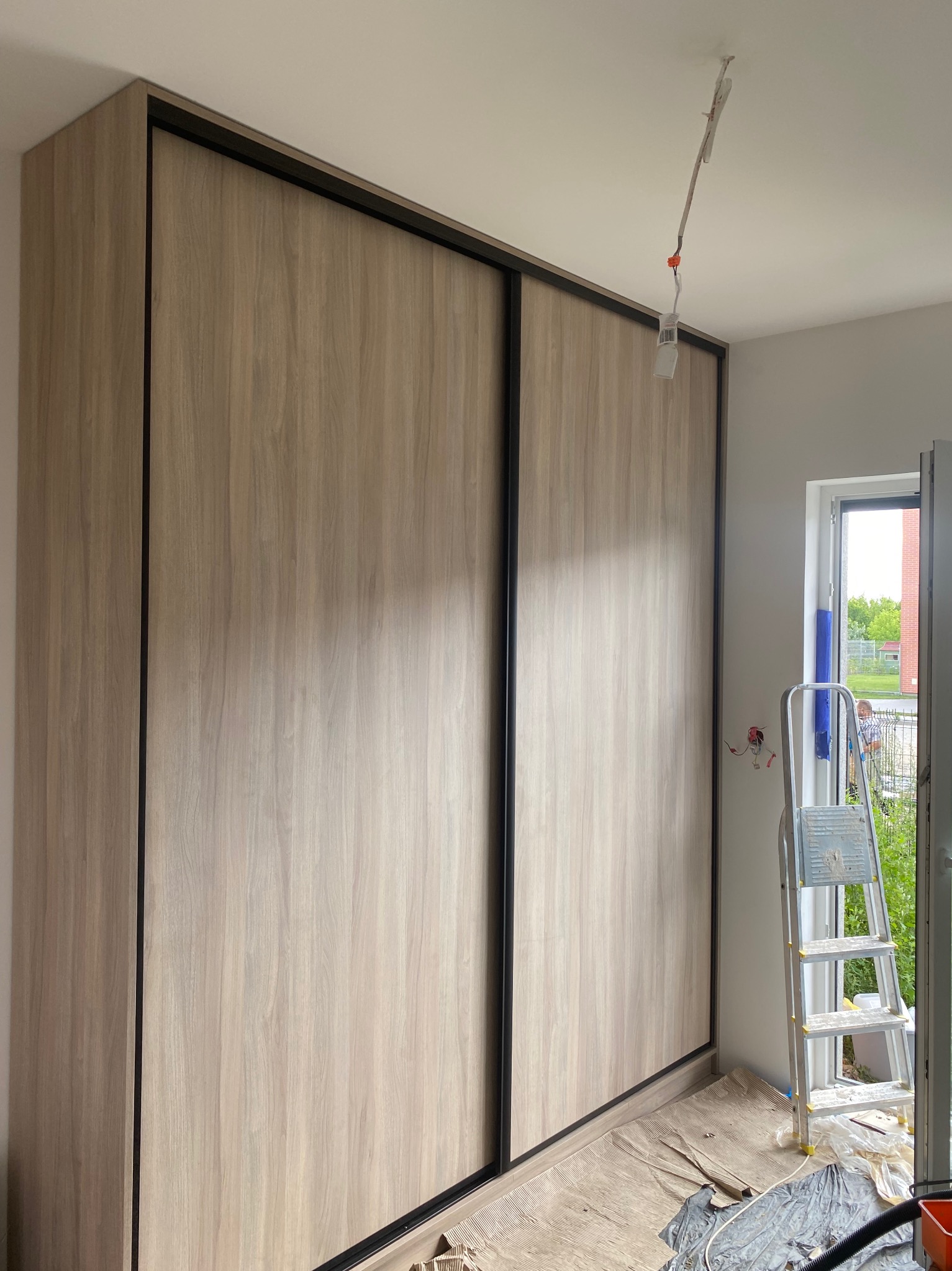 Jasna szafa przesuwna z drewnianymi frontami i czarnymi obramowaniami, widoczna drabina malarska, okno i fragment instalacji elektrycznej.