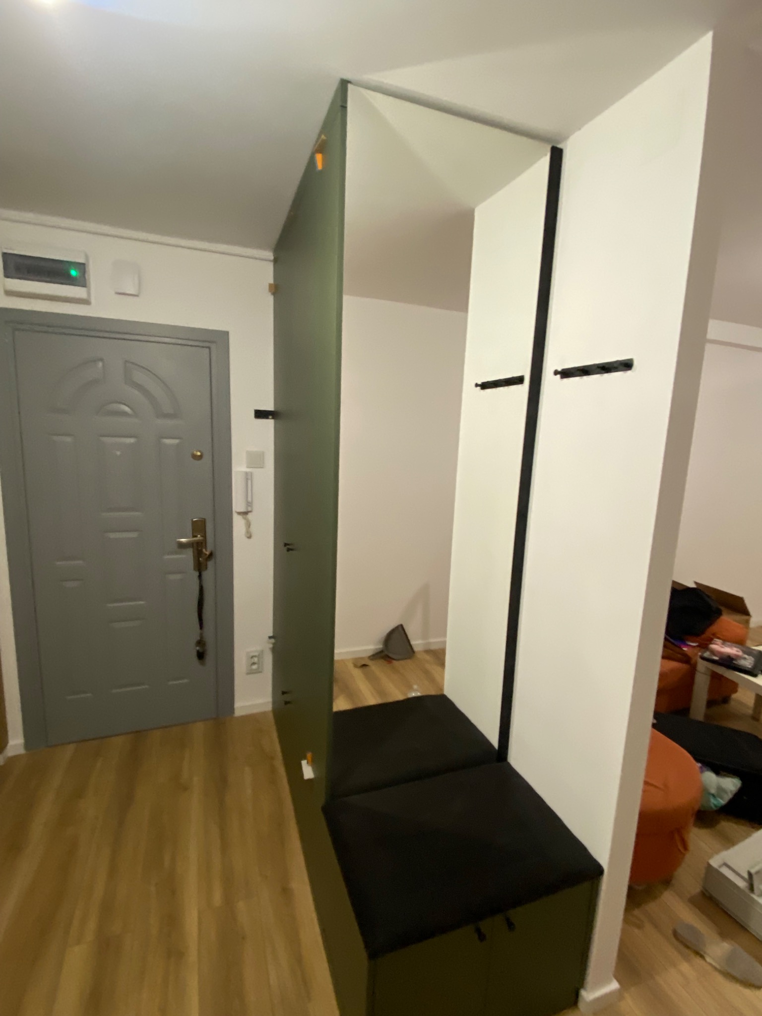 Nowoczesna szafa wnękowa z siedziskiem i wieszakami, w odcieniach bieli i zieleni, wkomponowana w przestrzeń przedpokoju. Minimalistyczny design z widocznymi detalami.