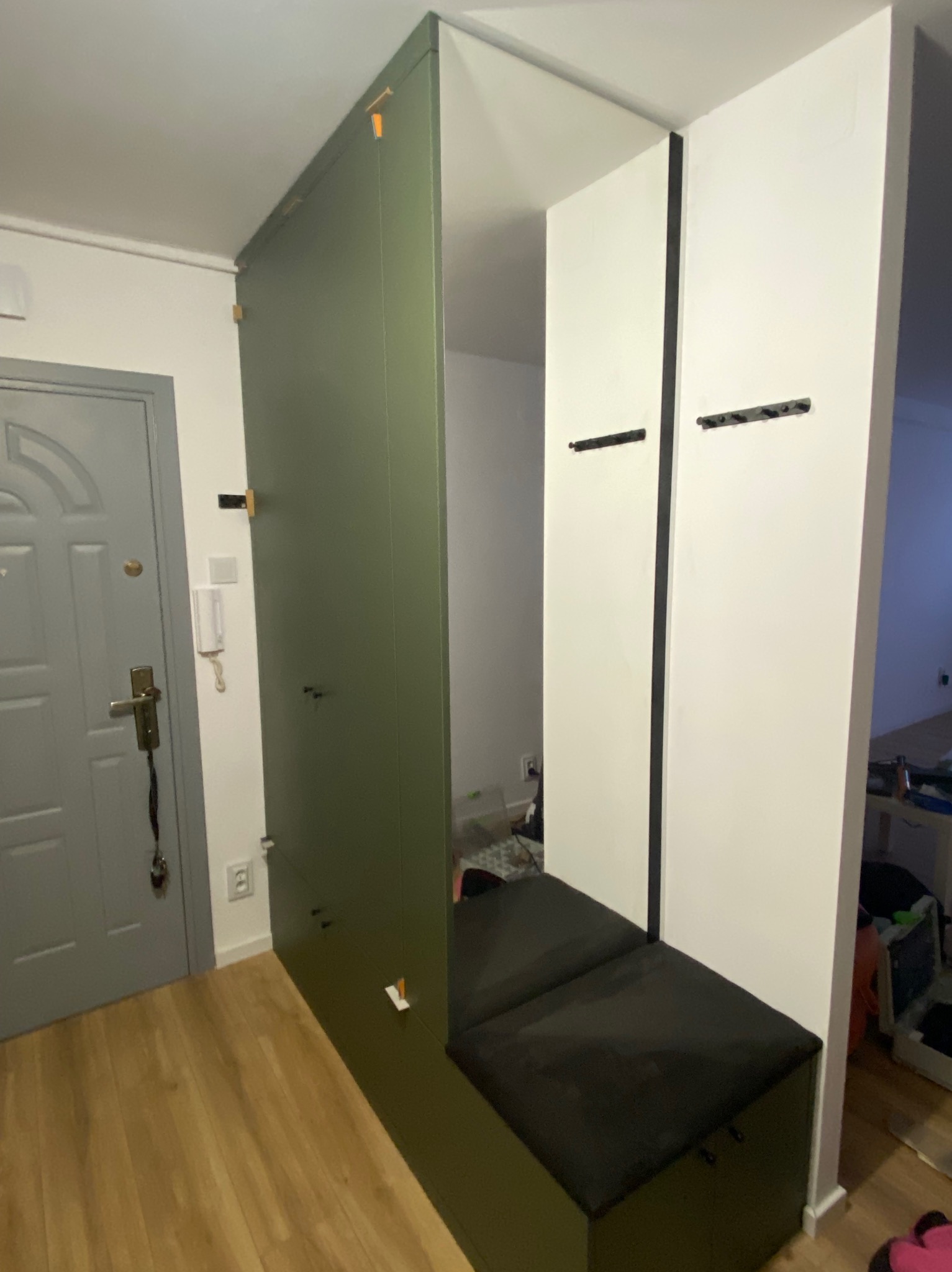 Funkcjonalna szafa w przedpokoju z siedziskiem i wieszakami, w odcieniach zieleni i bieli, z drewnianą podłogą i szarą ścianą z drzwiami.