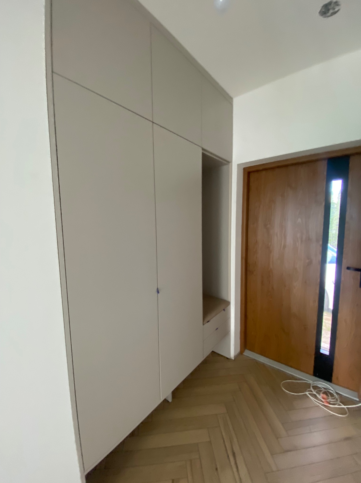 Jasna szafa wnękowa z siedziskiem w przedpokoju, drewniana podłoga w jodełkę, widoczne drzwi wejściowe z przeszkleniem, minimalistyczny design.