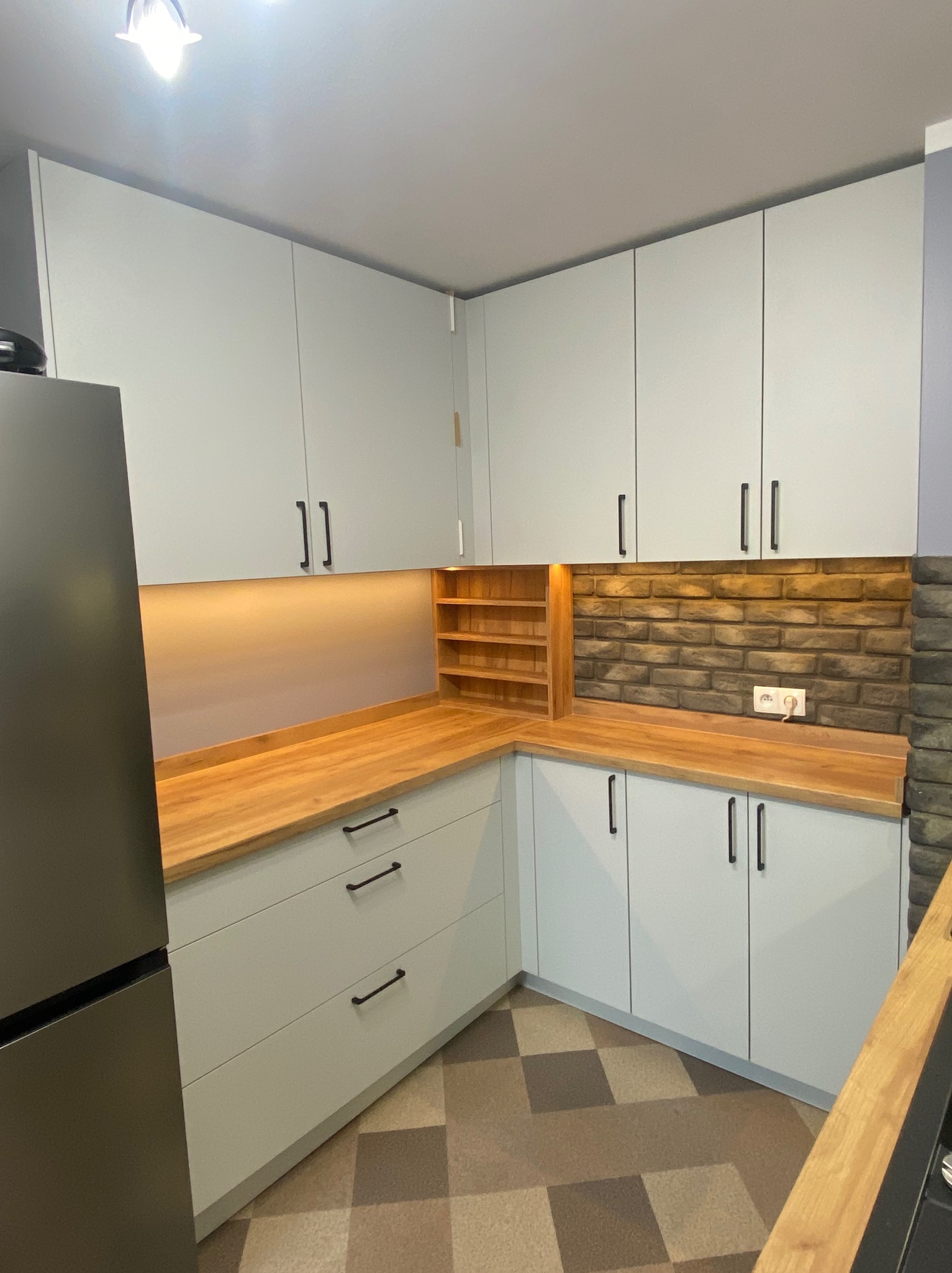 Jasna kuchnia z drewnianym blatem, oświetleniem podszafkowym i ceglaną ścianą. Widoczny fragment lodówki i podłoga w szachownicę.