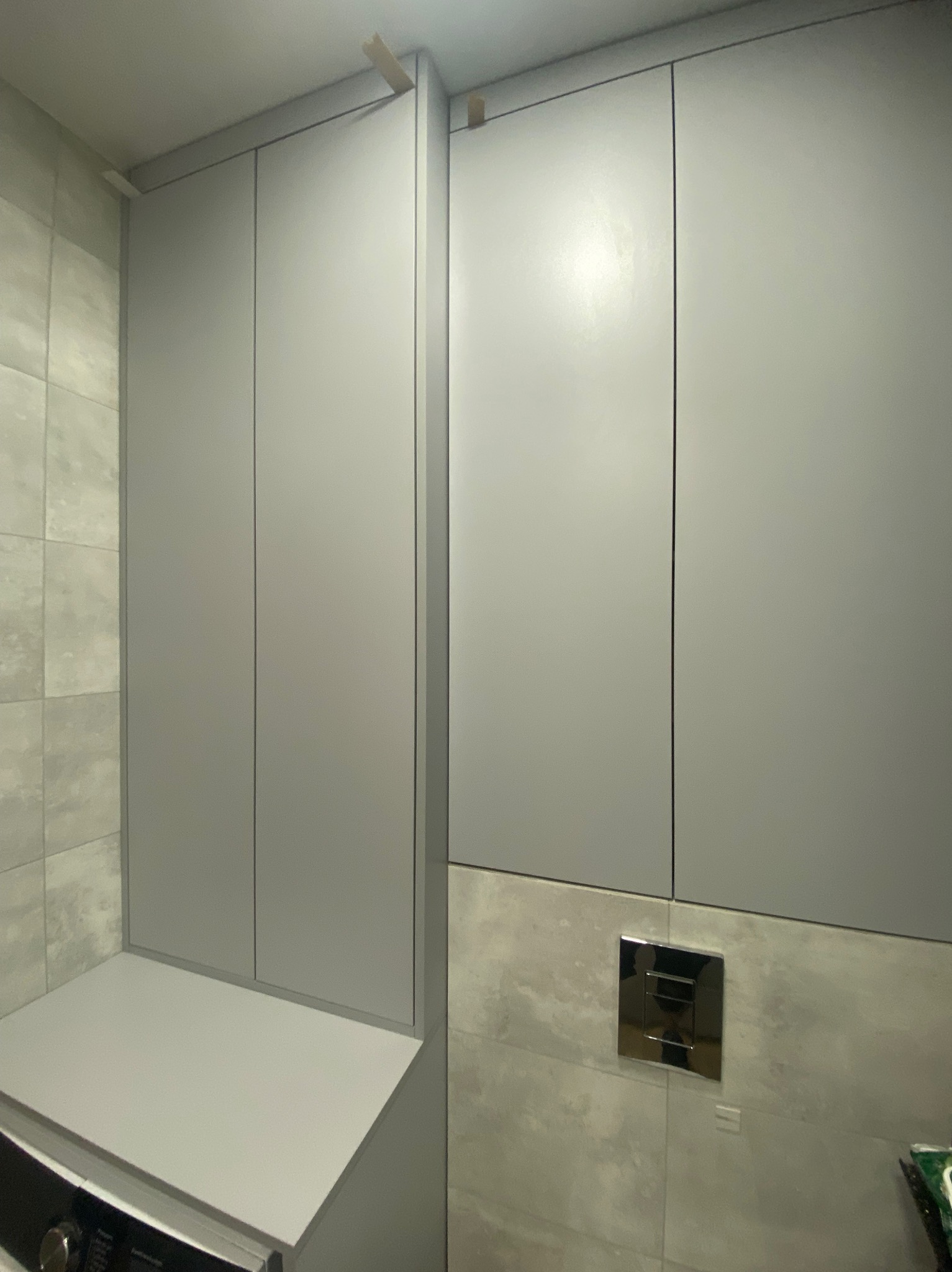 Szare szafki w łazience, zabudowa nad pralką i WC, z widocznymi elementami montażowymi. Minimalistyczny design, jasne płytki ceramiczne w tle.