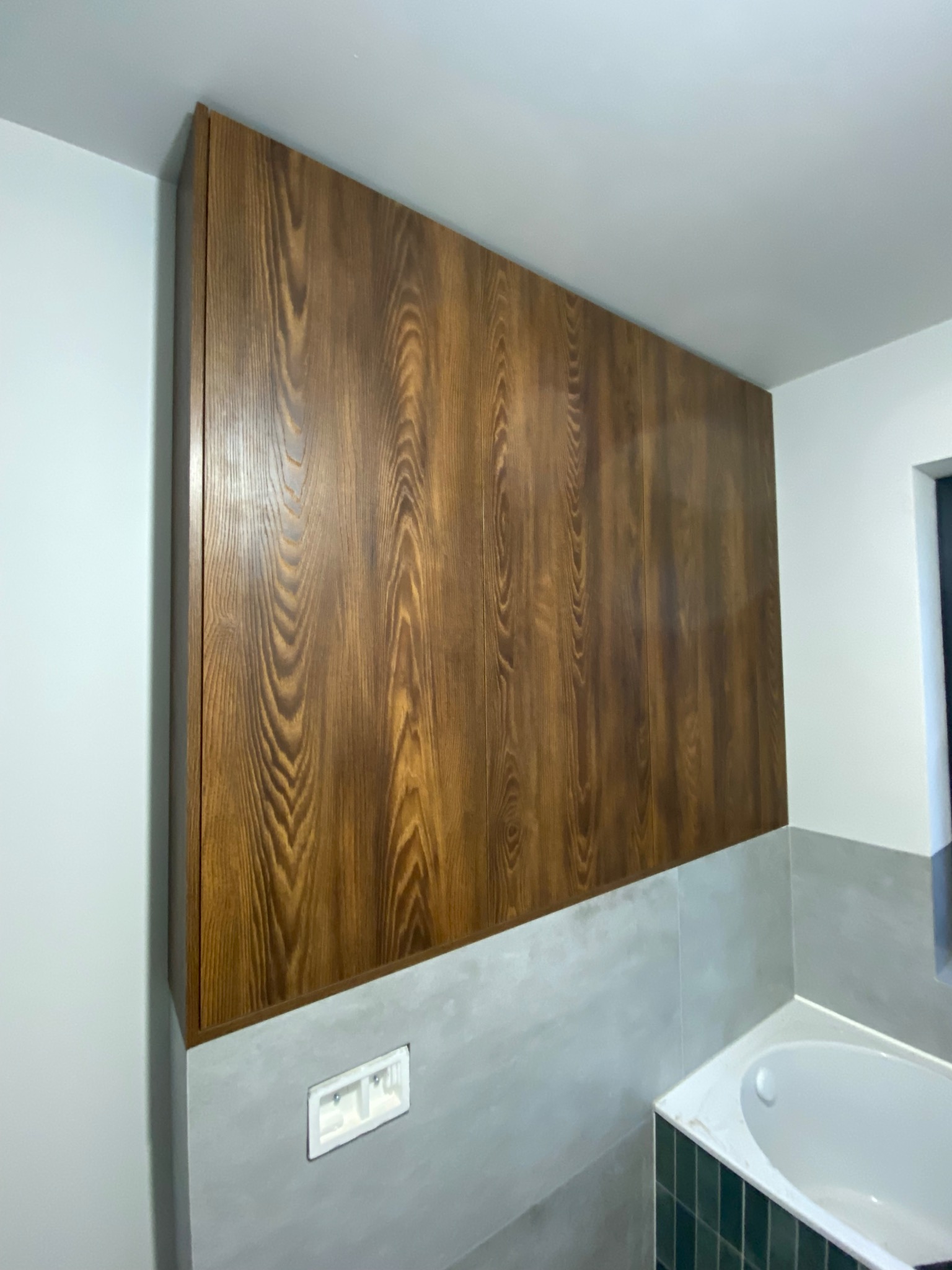 Drewniana szafa wnękowa z widocznym usłojeniem, wkomponowana w nowoczesną łazienkę z szarymi płytkami i białą wanną. Minimalistyczny design i ciepłe odcienie drewna.