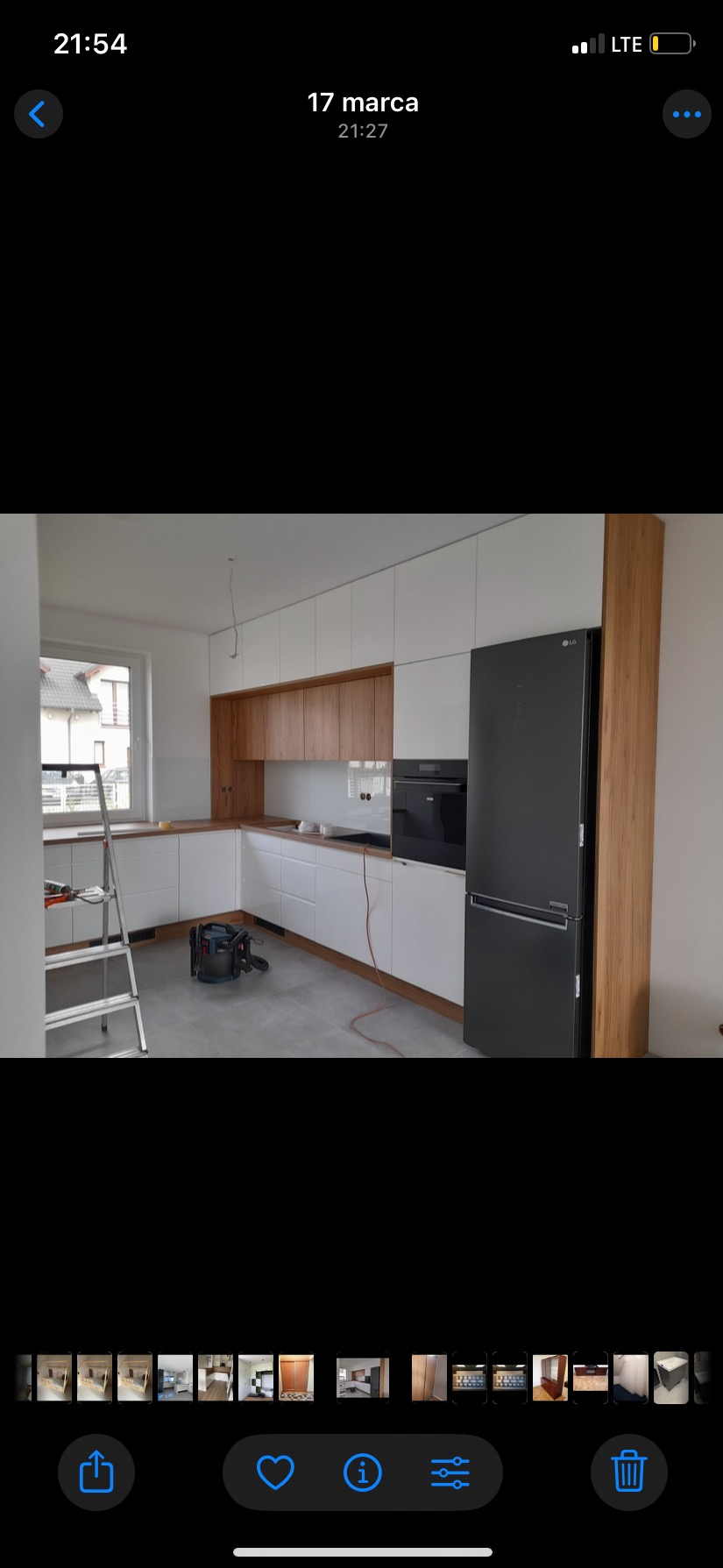 Nowoczesna, jasna kuchnia z białymi szafkami i drewnianymi akcentami. Widoczny sprzęt AGD oraz drabina i odkurzacz, sugerujące etap wykończenia.